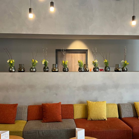 Ein modernes Interieur mit einer weißen Wand, einem grauen Sofa mit bunten Kissen und einer Holztür. Neun Vasen mit gelben Blumen stehen auf einer Leiste.