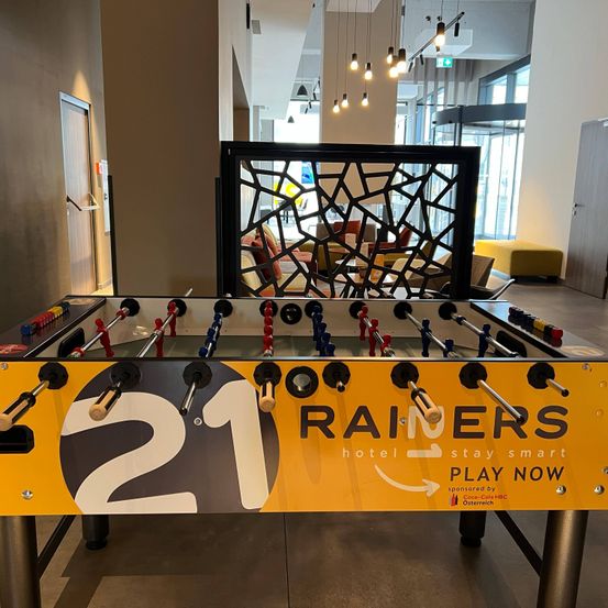 Ein Tischfußballtisch mit der Nummer 21 steht in einem Raum mit einer Glaswand. Der Raum hat einen gelben Ottoman und ein Metallgeländer.