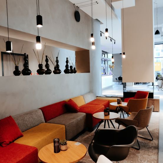 Eine moderne Lobby mit einem langen modularen Sofa, farbenfrohen Kissen und hängenden Lampen. Es gibt kleine runde Tische mit Stühlen und eine Glaswand, die das Innere reflektiert.