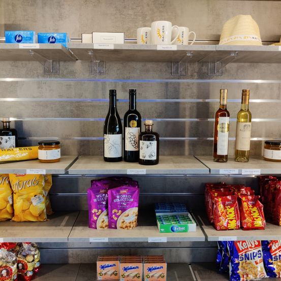 Ein Regal zeigt verschiedene Produkte, darunter Flaschen Wein, Snacks und andere Artikel. Flaschen Wein sind prominent ausgestellt, zusammen mit Gläsern und Snacktüten.