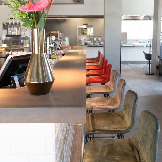 Innenansicht eines Restaurants mit einer Blumenvase auf einer Theke, Stühlen und Küchenbereich.
