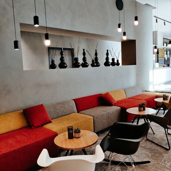 Eine moderne Lobby mit einer langen Couch, einigen Tischen und Stühlen. Die Couch hat rote und gelbe Kissen. Hängende Lampen und Regale mit schwarzen Vasen schmücken die Wand.