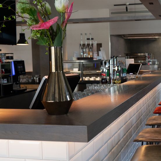 Eine Bartheke mit einer stilvollen Blumenvase in einem Restaurant. Die Theke ist aus Holz mit einer gefliesten Rückwand, und verschiedene Flaschen und Gläser sind ausgestellt.
