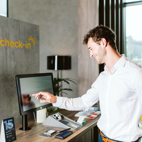Ein Mann steht vor einem Check-in-Monitor, lächelt und zeigt auf den Bildschirm. Hinter ihm zeigt ein Fenster eine Stadtlandschaft. Der Tresen hält verschiedene Büromaterialien.