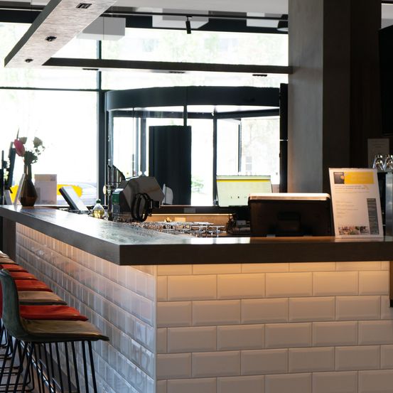 Ein heller, moderner Barbereich mit weißen Fliesenwänden und einer Theke mit Computer, Monitor und einer Vase mit Blumen.