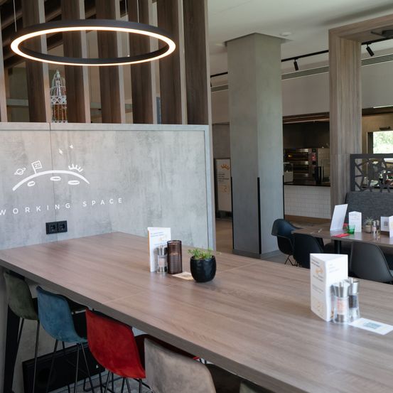 Ein Arbeitsbereich mit einem Holztisch und Stühlen. Eine Wand mit dem Text 'ARBEITSRAUM' und einer Illustration ist vorhanden. Eine Lampe hängt von der Decke ab.