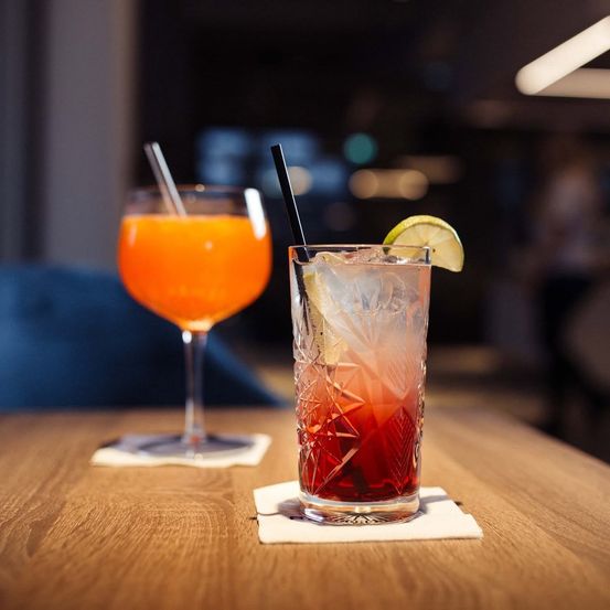 Zwei Cocktails auf einem Holztisch, einer mit roter Flüssigkeit und einer Zitronenscheibe, der andere mit orangefarbener Flüssigkeit und einem Strohhalm.