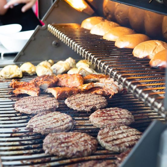 Eine Nahaufnahme eines Grills mit verschiedenen Fleischsorten, darunter Hamburger, Rippchen und Würstchen, mit Brötchen im oberen Rost.
