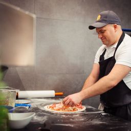 Ein Mann in einem weißen T-Shirt und einer schwarzen Schürze bereitet in einer Küche eine Pizza zu. Er fügt der Pizza auf einem Tresen Toppings hinzu.