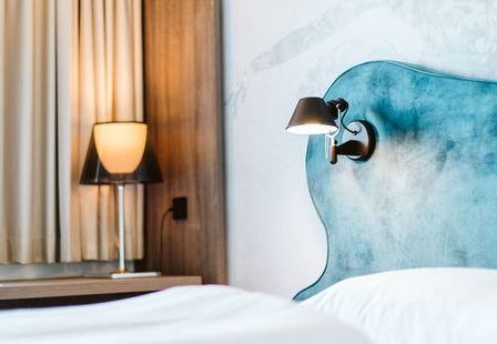 Ein Hotelzimmer mit blauem Kopfteil und weißer Bettwäsche. Eine Lampe steht auf dem Nachttisch, und eine weitere Leuchte ist an der Wand montiert.