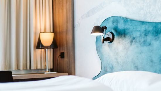 Ein Hotelzimmer mit blauem Kopfteil und weißer Bettwäsche. Eine Lampe steht auf dem Nachttisch, und eine weitere Leuchte ist an der Wand montiert.