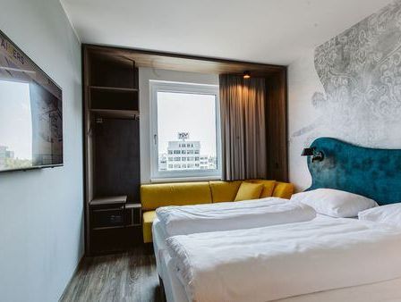 Ein modernes Hotelzimmer mit zwei Betten, einer gelben Couch und einem Fernseher an der Wand. Das Zimmer hat Blick auf ein Gebäude.