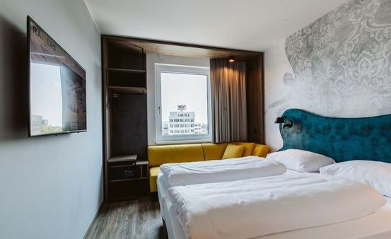 Ein modernes Hotelzimmer mit zwei Betten, einer gelben Couch und einem Fernseher an der Wand. Das Zimmer hat Blick auf ein Gebäude.