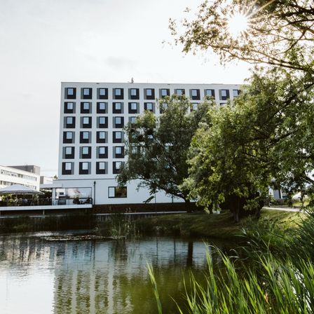 Ein großes weißes Hotel mit vielen Fenstern steht neben einem Teich mit Bäumen und Pflanzen. Die Sonne scheint hell am Himmel.