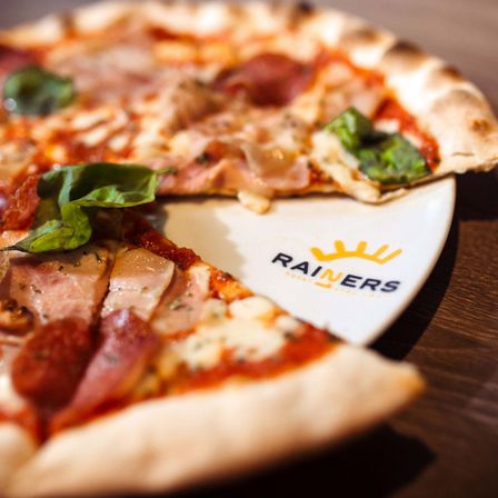 Ein Nahaufnahme eines Pizzastücks von Rainers mit Toppings wie Pepperoni, Schinken und Basilikum auf einem Holztisch.