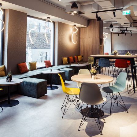 Ein moderner Indoor-Cafe-Bereich mit verschiedenen Tischen und Stühlen, einer mit Kissen belegten Bank und dekorativen Beleuchtungskörpern. Der Raum verfügt über große Fenster und ein klares, minimalistisches Design.