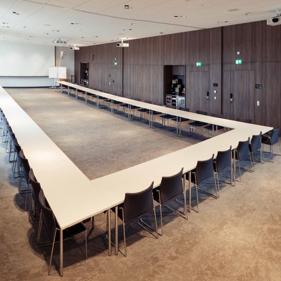 Ein leerer Konferenzraum mit einem langen Tisch und Stühlen. Der Raum hat Holzwände und eine weiße Decke. Es gibt zwei Whiteboards auf der linken Seite.
