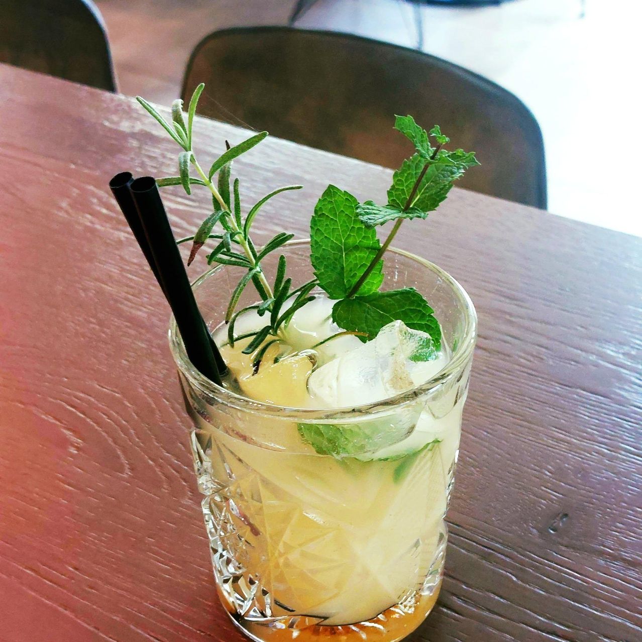 Ein Glas eines erfrischenden Cocktails mit Minzblättern, Rosmarin und schwarzen Strohhalmen auf einem Holztisch.