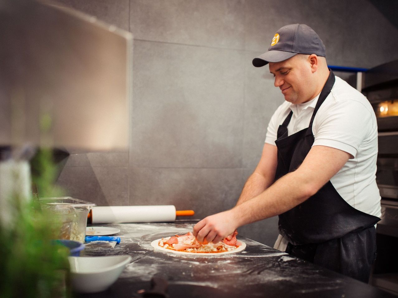 Ein Mann mit Hut und Schürze bereitet in einer Küche eine Pizza zu. Er fügt Toppings hinzu. Schüsseln und ein Nudelholz sind in der Nähe.