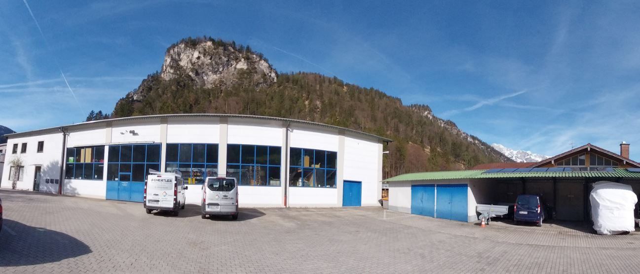 Ein weißes Gebäude mit blauen Türen steht vor einem Berg. Ein blauer Van ist unter einem grünen Dach geparkt.