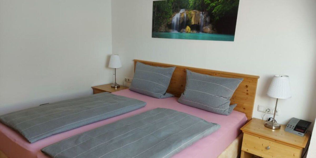 Ein Schlafzimmer mit einem Doppelbett, einem Holzkopfteil und einem an der Wand montierten Wasserfallbild. Ein Nachttisch hält eine Lampe, und ein weiterer Nachttisch steht auf der gegenüberliegenden Seite.