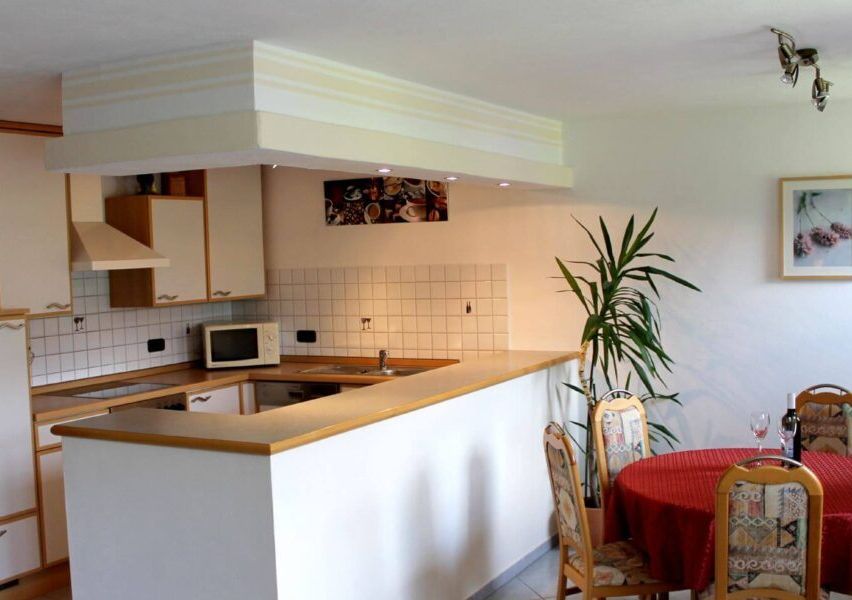 Eine Küche mit Mikrowelle, Spüle und Schränken. Ein Essbereich mit roter Tischdecke, Stühlen und einer Pflanze. Ein Bilderrahmen an der Wand.