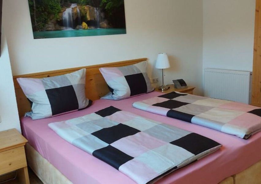 Ein Schlafzimmer mit einem Doppelbett, rosa Bettlaken und karierten Decken. Eine Lampe, ein Telefon und ein Heizkörper stehen auf dem Nachttisch. Ein Wasserfallbild hängt an der Wand.