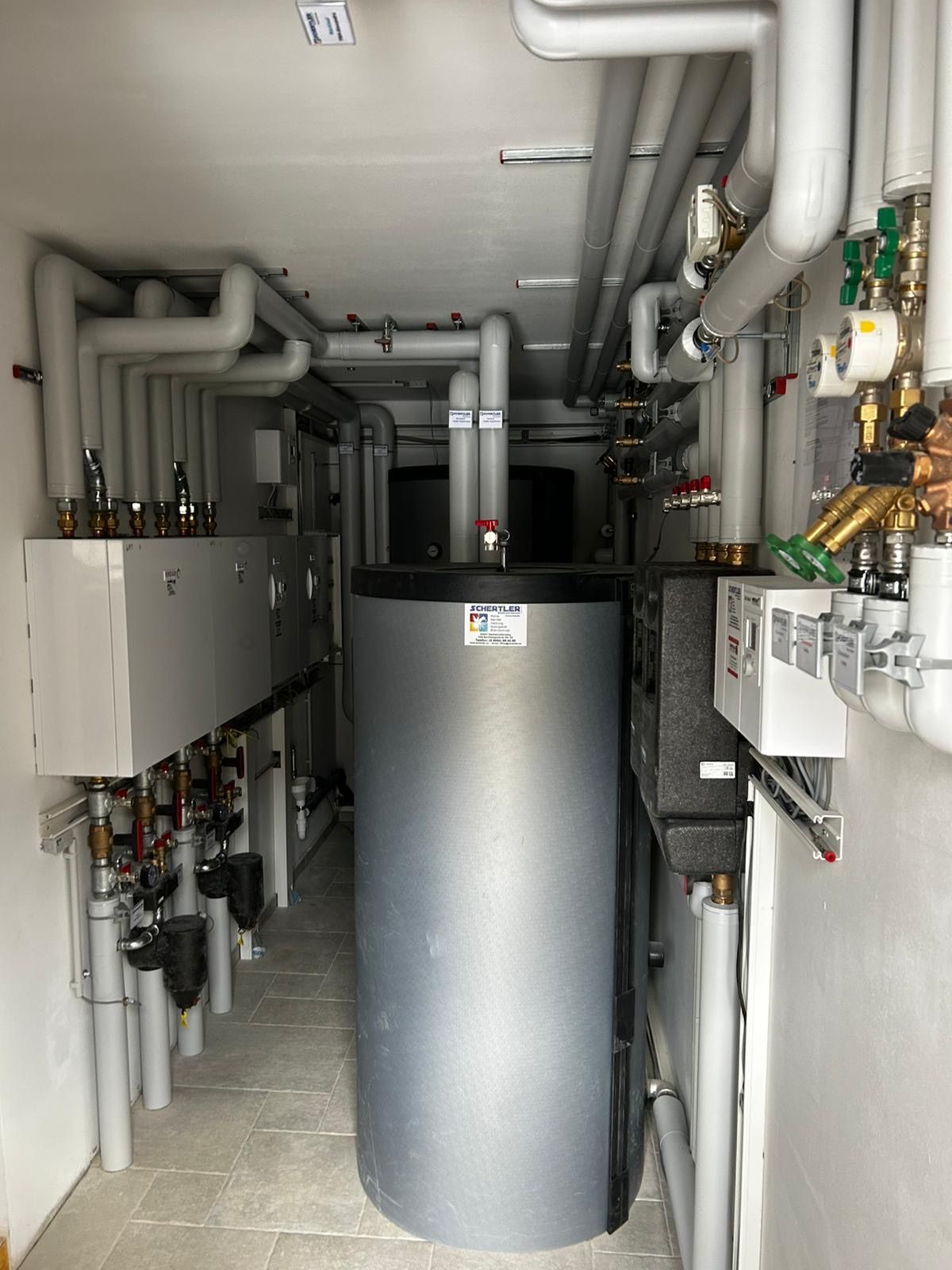 Ein Raum mit verschiedenen industriellen Geräten, darunter ein großer silberner Wassertank, der mit zahlreichen Rohren und Ventilen verbunden ist. Verschiedene Schalter und Bedienfeldern sind an den Wänden montiert.