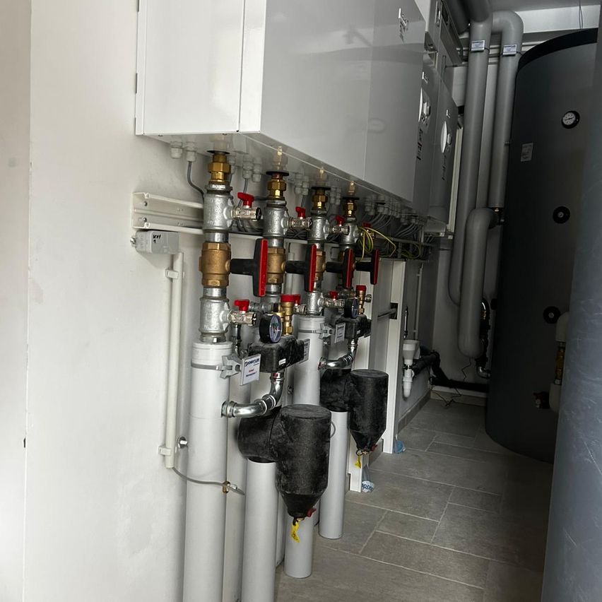 Ein Raum mit weißen Wänden enthält verschiedene Metallrohre, Messgeräte und Ventile. Ein großer Wassertank steht an der rechten Wand.