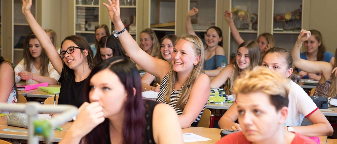Ein Klassenzimmer mit Schülern an Tischen. Eine Frau in einem schwarzen Shirt hebt die Hand, während eine andere Frau mit blonden Haaren in der Nähe sitzt, beide lächeln. Das Klassenzimmer hat Regale mit verschiedenen Gegenständen im Hintergrund.