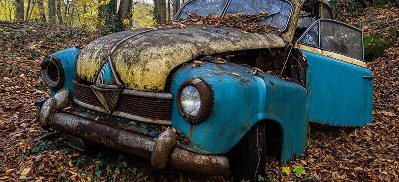 Ein altes blaues und gelbes Auto mit einem Nummernschild steht verlassen in einem Wald mit trockenen Blättern.