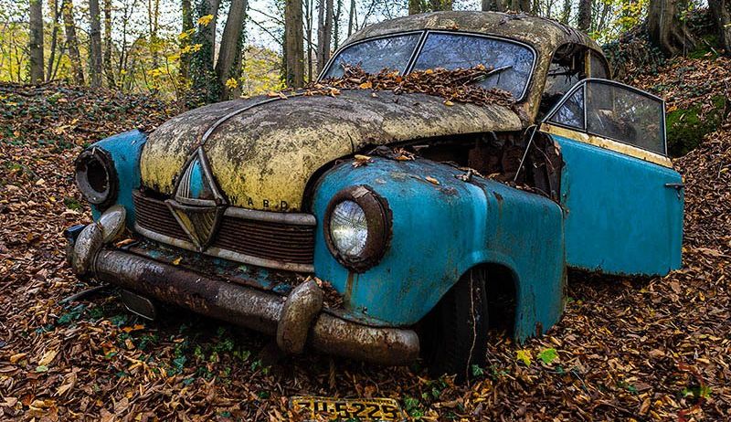 Ein altes blaues und gelbes Auto mit einem Nummernschild steht verlassen in einem Wald mit trockenen Blättern.