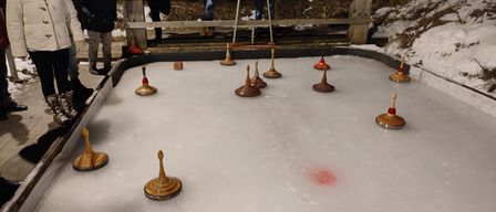 Eine Gruppe von Menschen beobachtet ein Spiel mit Drehtöpfen auf einer Eisbahn. Die Bahn hat Holzbarrieren und mehrere drehende Töpfe darauf.