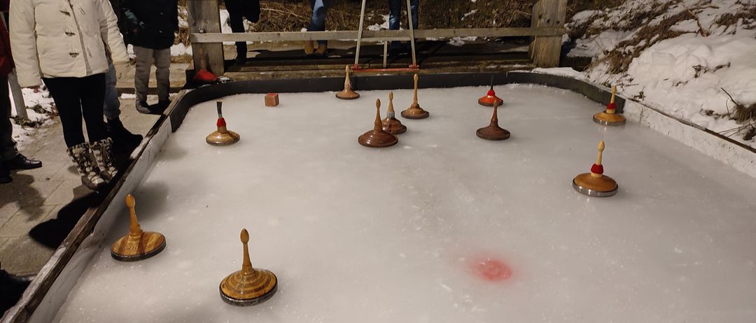 Eine Gruppe von Menschen beobachtet ein Spiel mit Drehtöpfen auf einer Eisbahn. Die Bahn hat Holzbarrieren und mehrere drehende Töpfe darauf.