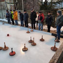 Eine Gruppe von Menschen in Winterkleidung beobachtet, wie jemand ein Kegelspiel auf einer vereisten Oberfläche spielt.