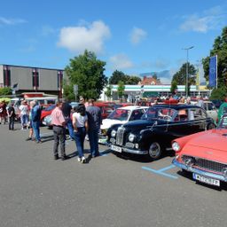 Eine Gruppe von Menschen versammelt sich um Oldtimer in einem Parkplatz unter einem klaren Himmel mit Wolken.