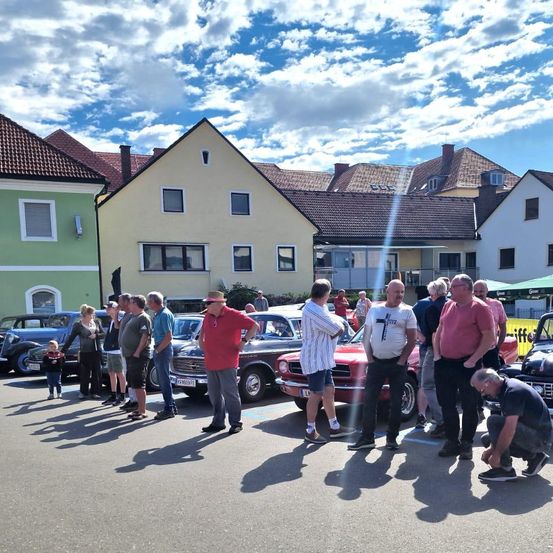 Eine Gruppe von Menschen steht um Oldtimer in einem Stadtplatz herum. Ein Mann hockt sich hin, um ein Auto zu inspizieren, während andere plaudern. Im Hintergrund befinden sich Gebäude und geparkte Fahrzeuge.