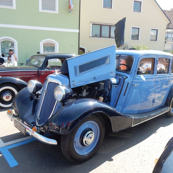Ein blaues Oldtimer-Auto mit offenem Motorhaube, geparkt vor einem Gebäude. Dahinter steht ein rotes Auto. Menschen stehen und gehen in der Nähe.