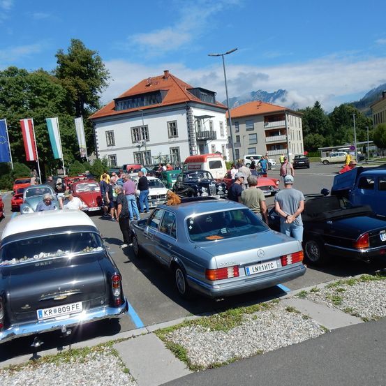 Oldtimer stehen auf der Straße vor einem Gebäude mit rotem Dach geparkt. Menschen gehen herum, möglicherweise bei einer Veranstaltung. Bäume und Flaggen sind in der Gegend sichtbar.