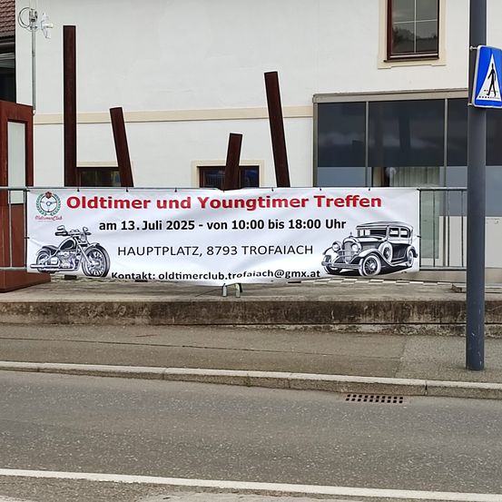 Ein Banner auf einem Geländer zeigt eine Veranstaltungsankündigung für Oldtimer und Youngtimer am 13. Juli 2025 von 10:00 bis 18:00 Uhr am Hauptplatz, 8793 Trofaiach. Es zeigt Abbildungen von Motorrädern und Autos.