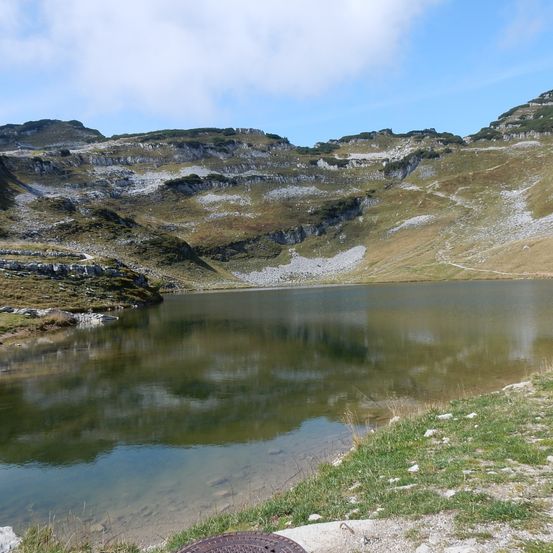 Ein ruhiger Bergsee mit der Reflexion des blauen Himmels und der Wolken. Felsige Klippen umgeben den See, und es gibt einen kleinen Grasfleck an der Seite.
