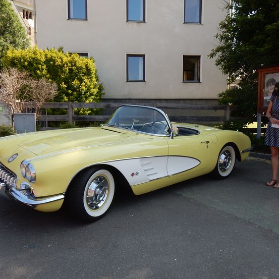 Ein gelb lackierter Corvette-Cabriolet-Oldtimer ist vor einem weißen Gebäude mit Glasfenstern und einem Holzzaun geparkt. Eine Frau steht in der Nähe.