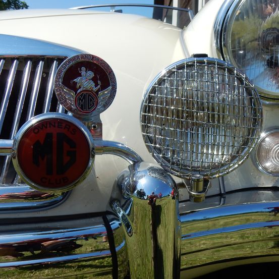 Nahaufnahme des vorderen Stoßfängers eines Oldtimers mit MG Owners Club-Emblem, Scheinwerfern mit Schutzgittern und Chrom-Details.