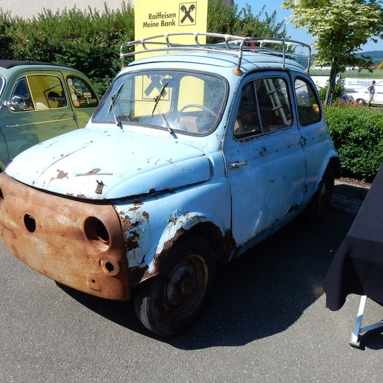 Ein alter blauer Fiat 500 mit Rostflecken und Dachträger steht neben einem gelben Schild.