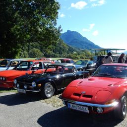 Ein rotes und schwarzes Auto ist unter einem Baum geparkt. Ein rotes Auto ist dahinter geparkt. Ein Baldachin ist in der Nähe der Autos. Berge sind im Hintergrund.