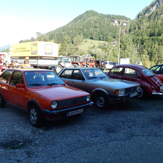 Mehrere Autos sind in einem Außenbereich geparkt. Ein roter Volkswagen Golf und ein grauer Ford stehen vorne. Dahinter befinden sich mehrere Fahrzeuge, darunter Lastwagen und Traktoren. Ein Berg ist im Hintergrund.