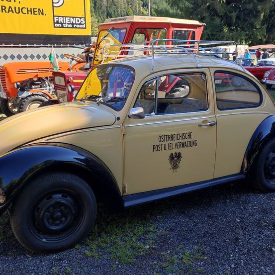 Ein Vintage Volkswagen Käfer mit Gepäckträger steht auf Kies geparkt, in der Nähe eines gelben Trucks mit Friends on the Road Schild.