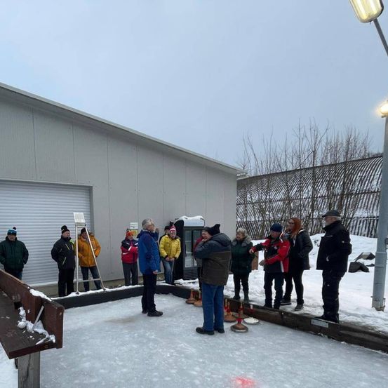 Eine Gruppe von Menschen in Winterkleidung versammelt sich auf einer Eisbahn und führt ein Gespräch. Im Hintergrund befindet sich ein Gebäude mit einer Garagentür.