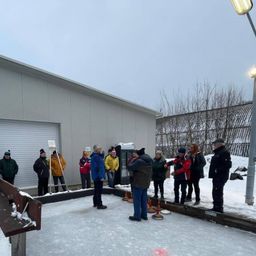 Eine Gruppe von Menschen in Winterkleidung versammelt sich auf einer Eisbahn und führt ein Gespräch. Im Hintergrund befindet sich ein Gebäude mit einer Garagentür.