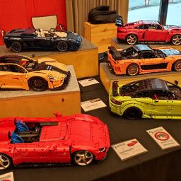 Eine Ausstellung verschiedener Lego-Sportwagen in verschiedenen Farben auf Holzregalen und einem Tisch. Es gibt Schilder, die darauf hinweisen, nicht anzufassen.
